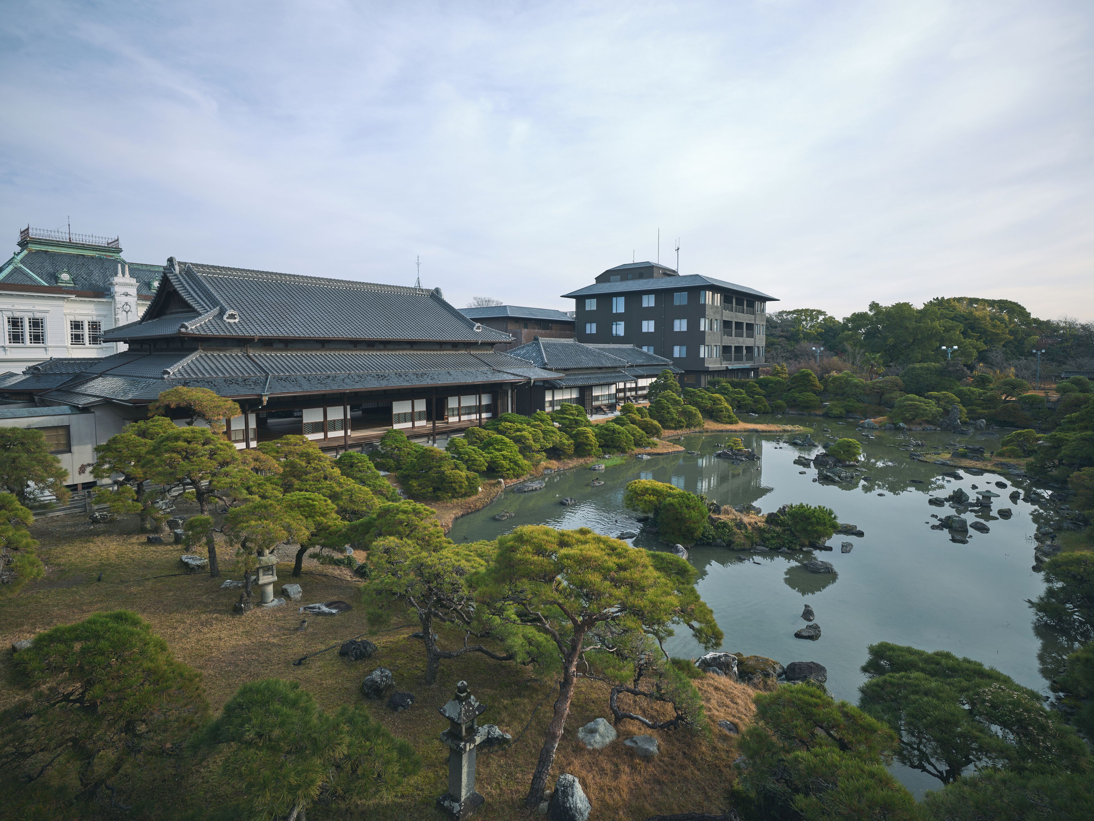 A traditional Japanese building complex with multiple structures, a pond, and surrounding trees.