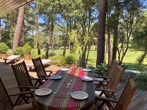 Uma área de refeições ao ar livre coberta, com uma mesa posta para quatro pessoas, rodeada por cadeiras de madeira, com vista para um jardim exuberante e um campo de golfe.