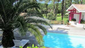 Uma área de piscina com um deck de madeira, uma palmeira e um pequeno edifício com telhado vermelho.