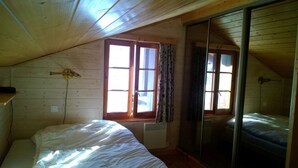 Een slaapkamer met een schuine kap, houten wanden, een bed, een raam met gordijnen en een spiegel.