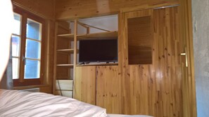 Une pièce en bois avec un lit, un téléviseur fixé sur une étagère en bois et une fenêtre avec des stores.