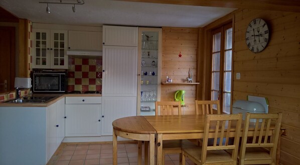 Une cuisine avec des meubles blancs, un four à micro-ondes, une table à manger en bois avec des chaises et une horloge au mur.