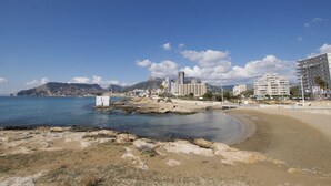 A coastal cityscape with a sandy beach, a rocky shoreline, and several multi-story buildings in the background. - A110 EDIFICIO LA REINA 8º 45 (Calpe)