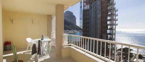 A balcony with a view of the sea and a mountain, a potted plant, and a table with chairs.