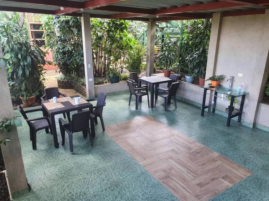 Un patio couvert, aménagé avec des tables et des chaises, entouré de plantes en pot et offrant une vue sur le jardin.