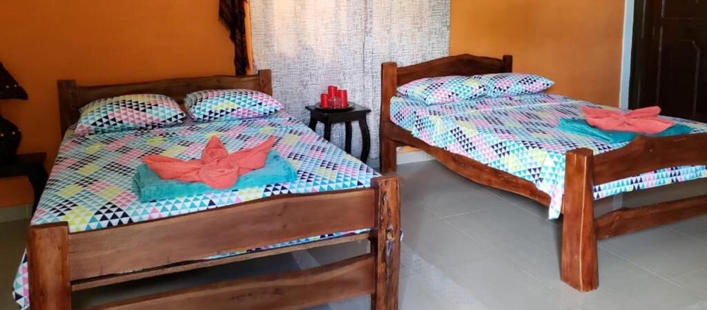 Due letti di legno con coperte e asciugamani colorati, un tavolino con bicchieri rossi e una tenda sullo sfondo.