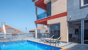 Un área de piscina en la azotea con una piscina azul cristalina, sillas de descanso y un edificio moderno con puertas de vidrio y un balcón.