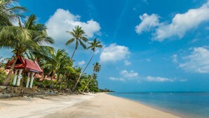 A beach with a red-roofed structure, palm trees, and clear blue skies. - Magical, 2 Bedroom Apartment in Beachside Complex (Koh Samui)