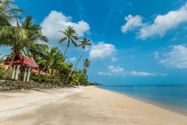 A beach with a red-roofed structure, palm trees, and clear blue skies. - Magical, 2 Bedroom Apartment in Beachside Complex (Koh Samui)