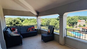 Un balcon abrité avec un canapé et un fauteuil, une piscine en arrière-plan et une vue sur les arbres et les bâtiments.