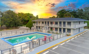 A two-story motel with a swimming pool, a parking lot, and a view of the sunset.
