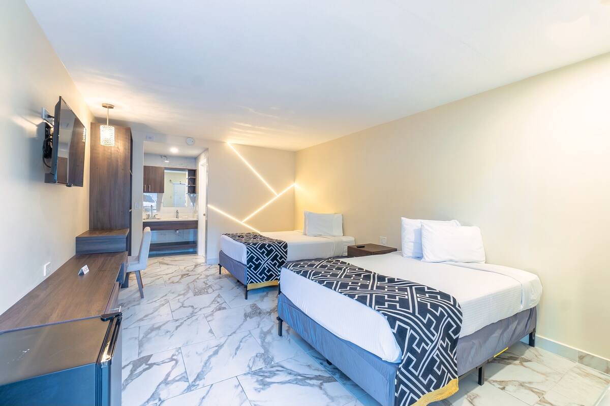 Hotellihuone, jossa on kaksi sänkyä, työpöytä, peili ja kylpyhuone, joka näkyy avoimesta ovesta.