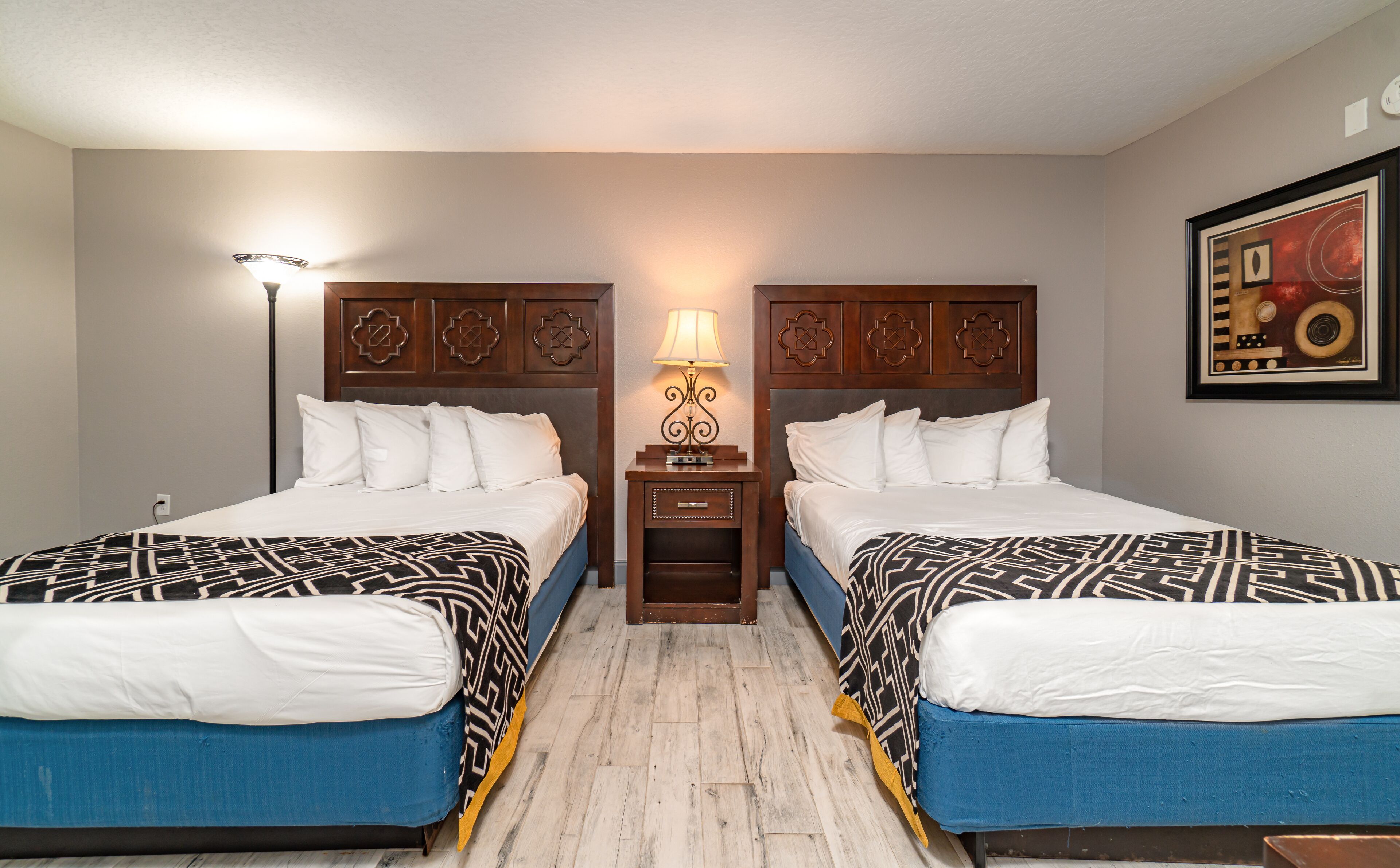 A hotel room with two beds, a nightstand with a lamp, a framed picture on the wall, and a wooden floor.