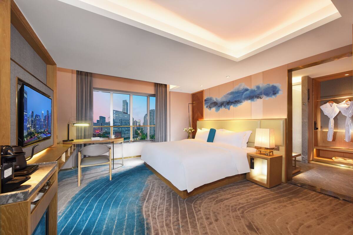 A modern hotel room with a large bed, a desk, a TV, and a city view.