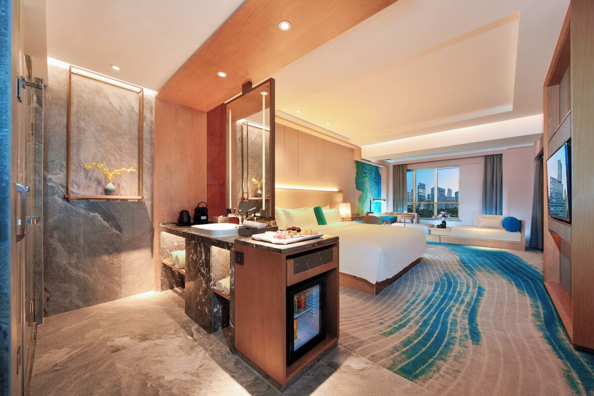A modern hotel room with a marble bathroom, a large bed, a sitting area with a view, and a small kitchenette.