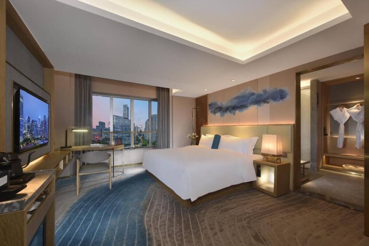 A hotel room with a large bed, a desk, a TV, and a view of the cityscape.