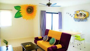 Ein Wohnzimmer mit einem violett-gelben Sofa, einem Deckenventilator, einer großen Sonnenblumen-Dekoration und einem Essbereich mit Tisch und Stühlen.
