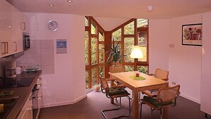 A dining area with a wooden table, chairs, and a lamp. There is a window with a view of greenery outside.