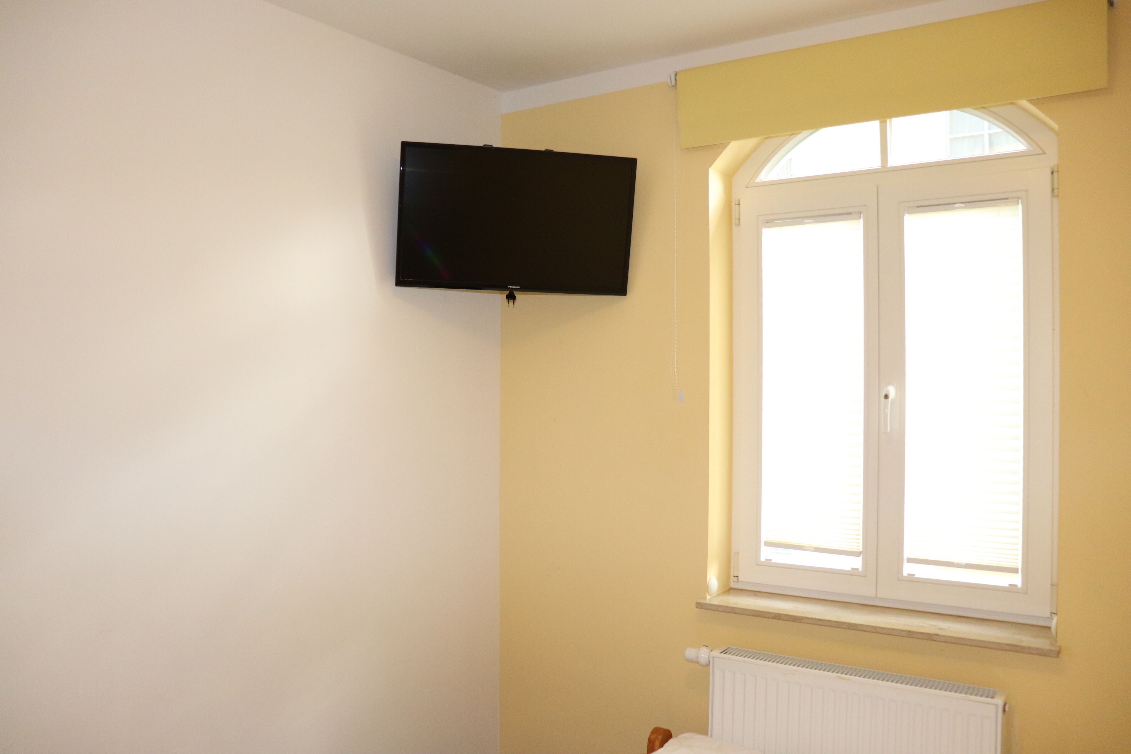 A room with a mounted TV, a window with blinds, and a radiator.