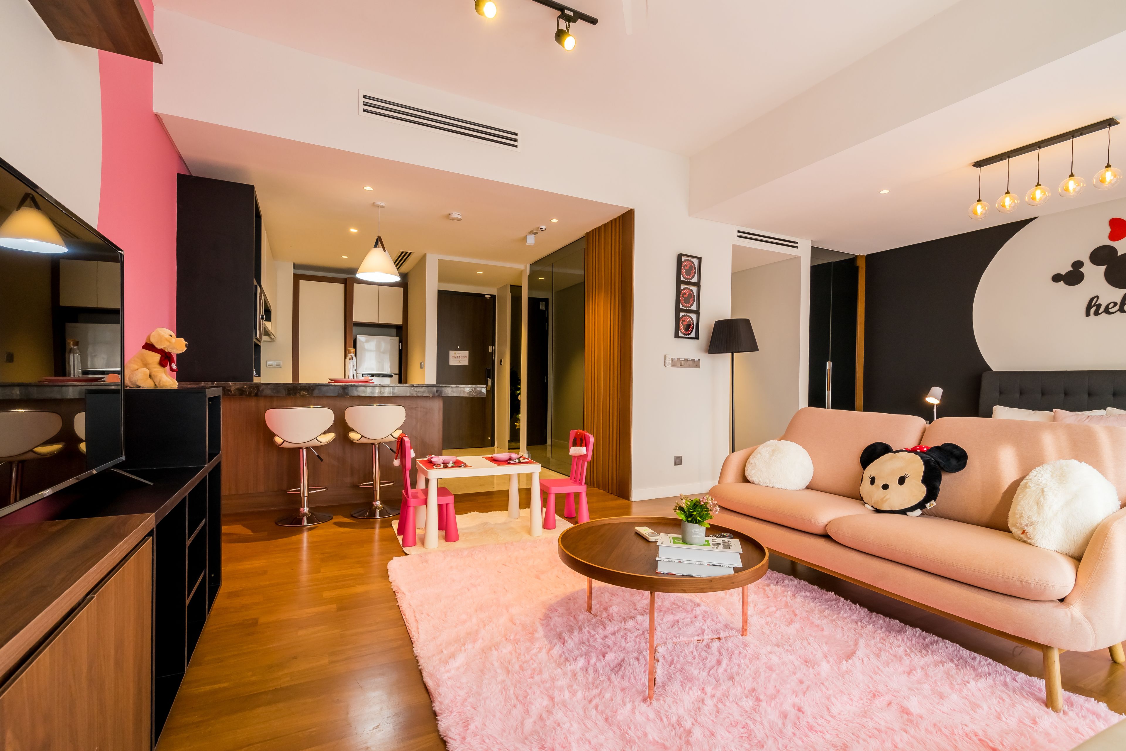 A modern living room with a pink sofa, a coffee table, and a pink rug. There is a kitchen area with a bar counter and stools.
