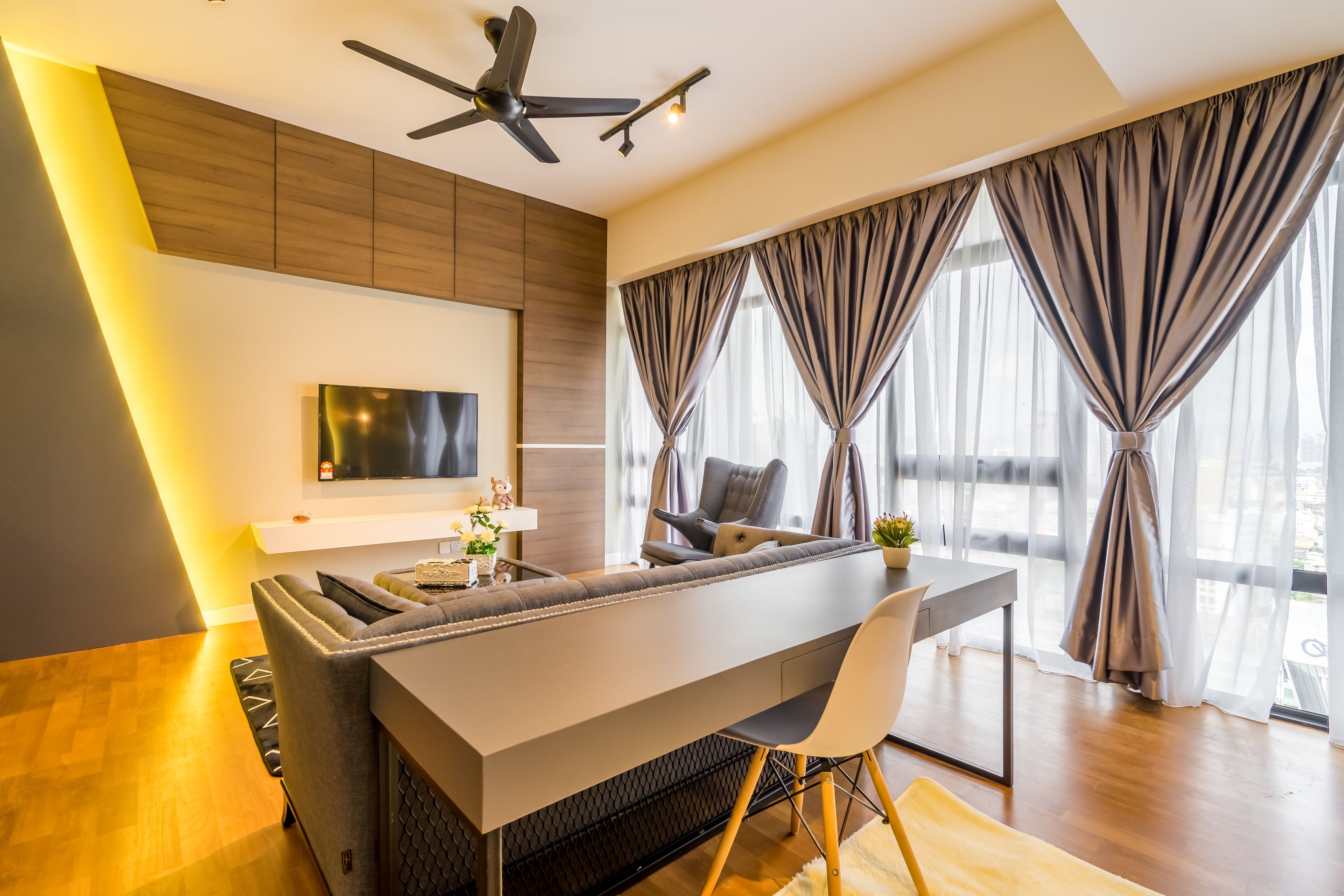 A modern living space with a central island, a ceiling fan, a flat-screen TV, and large windows with curtains.