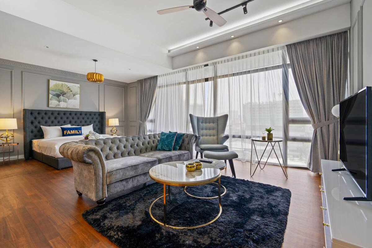 Une chambre d’hôtel moderne équipée d’un lit, d’un canapé, d’un fauteuil, d’une télévision et d’une grande fenêtre avec des rideaux.