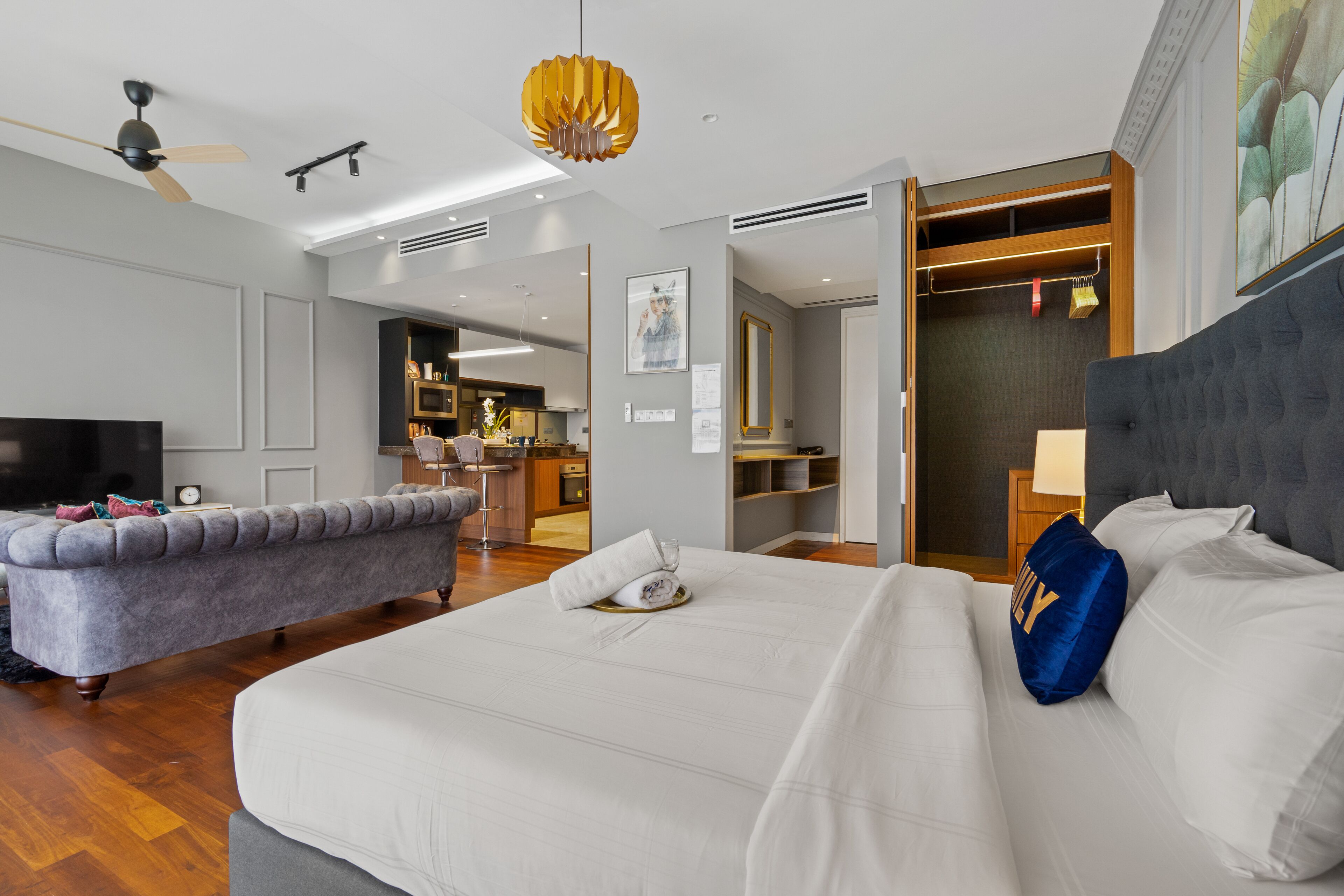 Quarto de hotel com cama, sofá, televisão, ventoinha no teto e área de cozinha visível através de uma porta aberta.