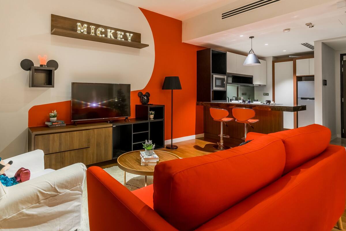 Un salon moderne avec un canapé rouge, un téléviseur, une petite table avec une plante, et un coin cuisine à l’arrière-plan.