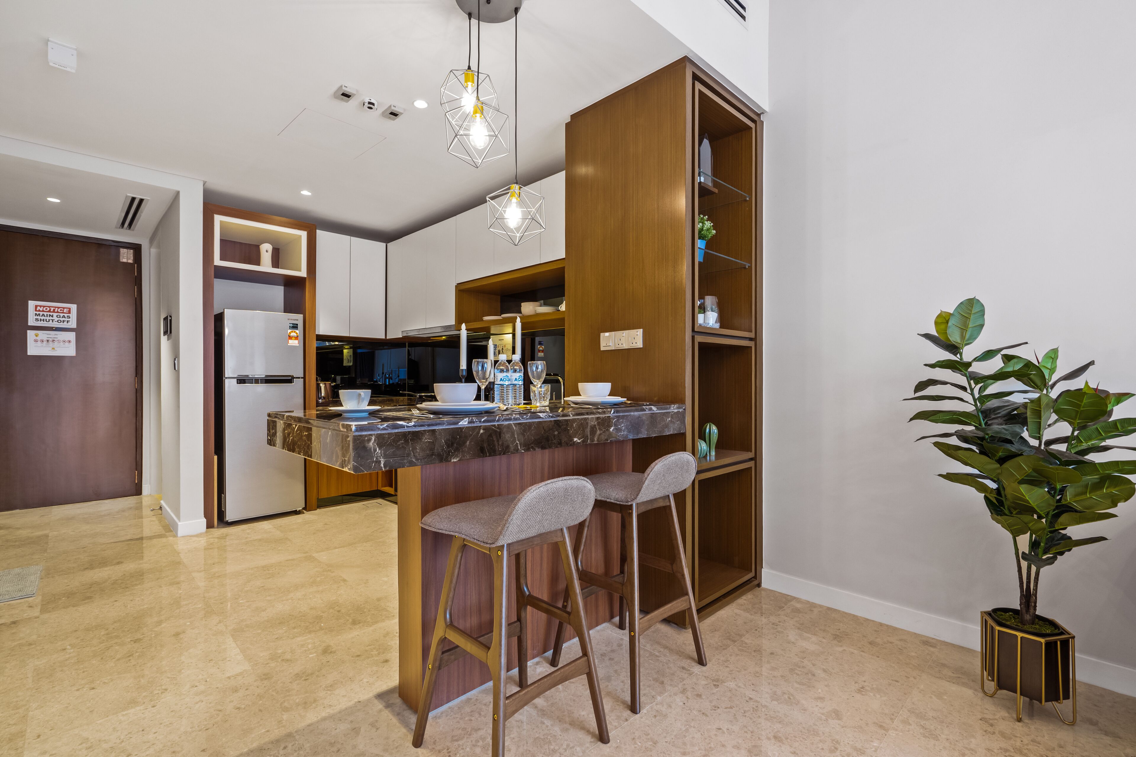 A modern kitchen with a marble countertop, two bar stools, a built-in bookshelf, and a potted plant.