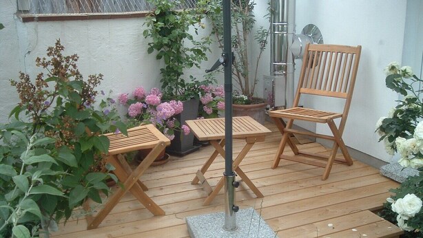 Eine Holzterrasse mit zwei Holzstühlen und einem Tisch, umgeben von Blumentöpfen und Blumen.