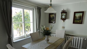 Ein Esszimmer mit Holztisch, weißen Stühlen, einer Blumenvase, einer Wanduhr und einem Fenster mit Vorhängen.