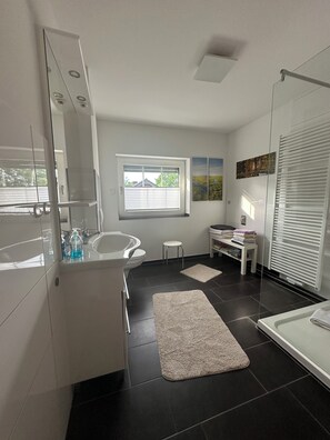 Et moderne badeværelse med et stort spejl, et badekar, et toilet og en håndvask. Der er et vindue med skodder, en lille skammel og en bøjle til håndklæder.