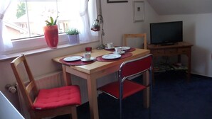 A small, cozy room with a wooden table set for two, two red chairs, a television on a wooden stand, and a window with a view of the outside. - NATURE and PEACE 🍀 Cozy, quiet 2-room vacation apartment in Münster (Münster)