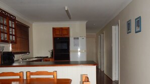 A kitchen with wooden cabinets, a refrigerator, and a dining area with a table and chairs. - Manly Peninsular (Wynnum)