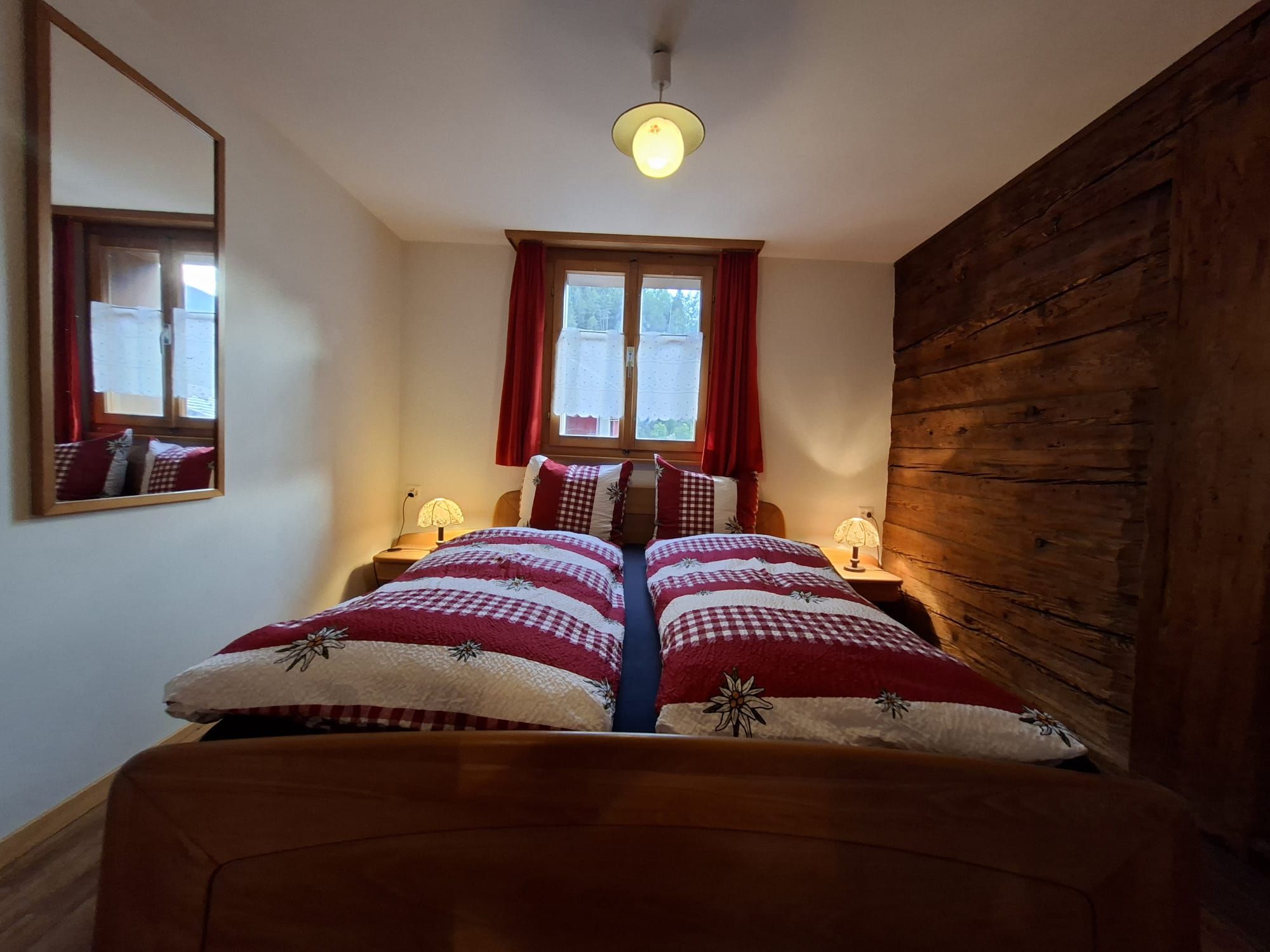 A bedroom with a wooden headboard, a bed with a checkered bedspread, two bedside lamps, and a window with red curtains.