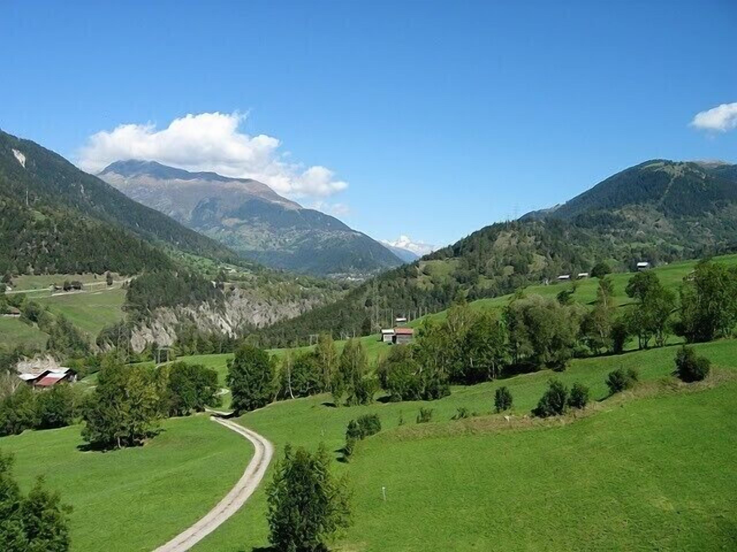 A rural landscape with a winding road, green fields, scattered trees, and a few buildings.
