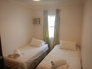 A hotel room with two single beds, each with a rolled towel, a window with green curtains, and an air conditioning unit on the wall.