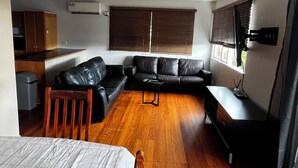 A compact living space with a sofa, a small table, a wooden chair, a bed, a kitchenette, and a large window with blinds. - Cedar Lookout (Wynnum)