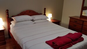 A neatly made bed with white bedding and a red towel, wooden nightstands with lamps, and a wooden dresser with drawers. - Cedar Lookout (Wynnum)