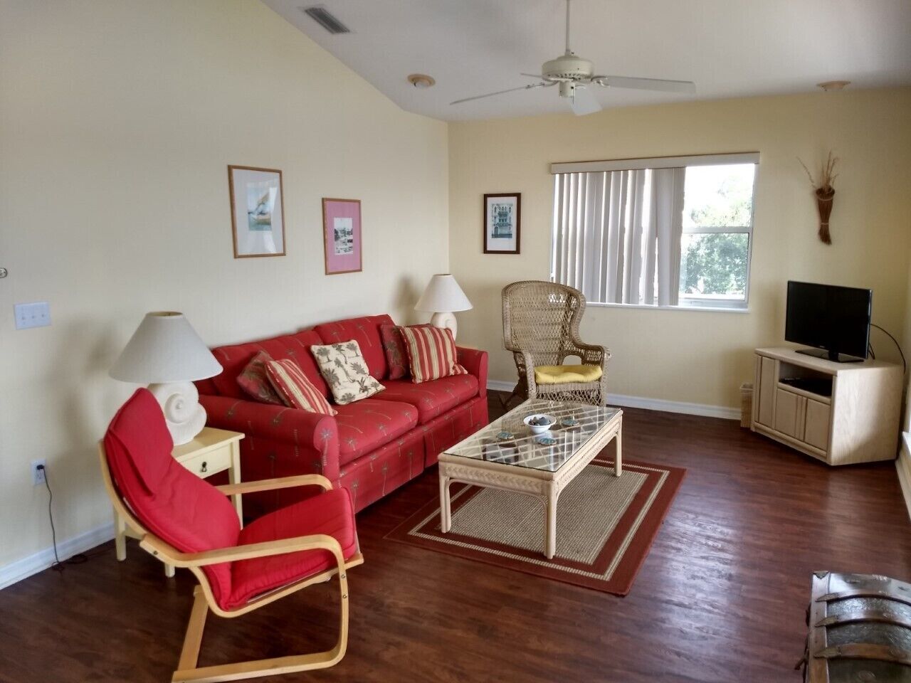 A living room with a red sofa, a glass coffee table, a wicker chair, a TV stand, and a ceiling fan.