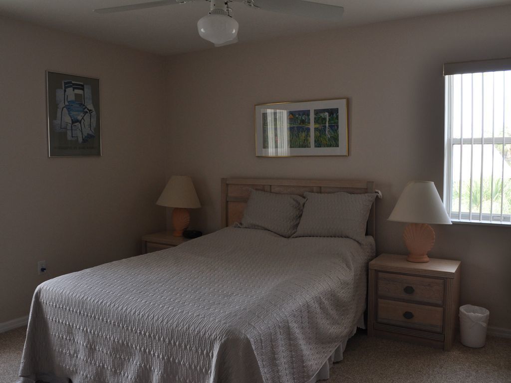 A bedroom with a bed, two bedside tables with lamps, a ceiling fan, a framed picture on the wall, and a window with blinds.