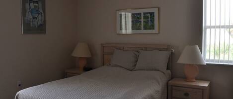 A bedroom with a bed, two bedside tables with lamps, a ceiling fan, a framed picture on the wall, and a window with blinds.