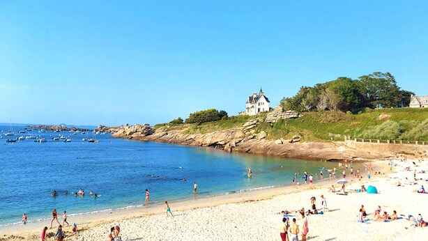 Una playa con gente, barcos y un edificio parecido a un castillo en una costa rocosa.