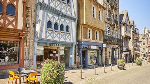 Calle con arquitectura europea tradicional, incluyendo edificios con entramado de madera y una tienda con el letrero 'JE BELLIE CHAUSEUR'.