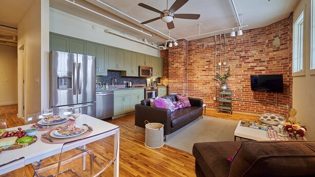 A modern kitchen with stainless steel appliances, a dining area with a glass table and chairs, a living room with a sofa and a TV mounted on a brick wall. - Luxury Loft in Downtown Asheville: chic, central, in the middle of all the fun! (Asheville)