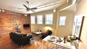 A modern living room with a brick wall, wooden flooring, a ceiling fan, a flat-screen TV mounted on the wall, and a dining area with a glass table and chairs. - Luxury Loft in Downtown Asheville: chic, central, in the middle of all the fun! (Asheville)
