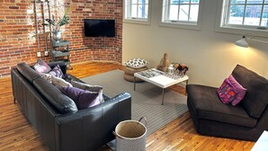 A living room with a brick wall, wooden flooring, a black leather sofa, a white coffee table, and a brown armchair. - Luxury Loft in Downtown Asheville: chic, central, in the middle of all the fun! (Asheville)