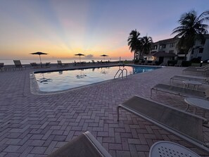 Ein Poolbereich mit Liegestühlen und Sonnenschirmen, Blick auf das Meer und ein Sonnenuntergang im Hintergrund.