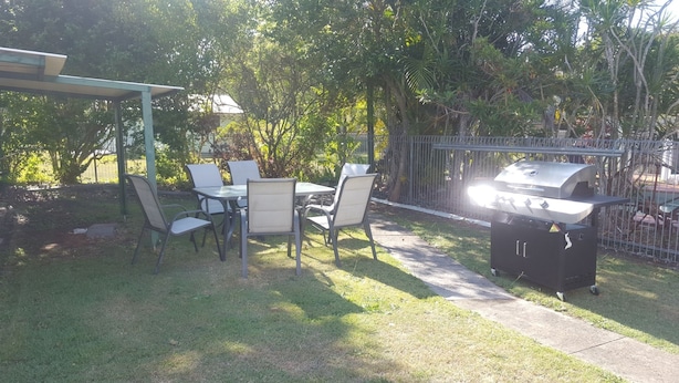 A backyard with a covered patio table and chairs, a barbecue grill, and a fenced area with trees. - Sailor's Rest Holiday House (Lota)