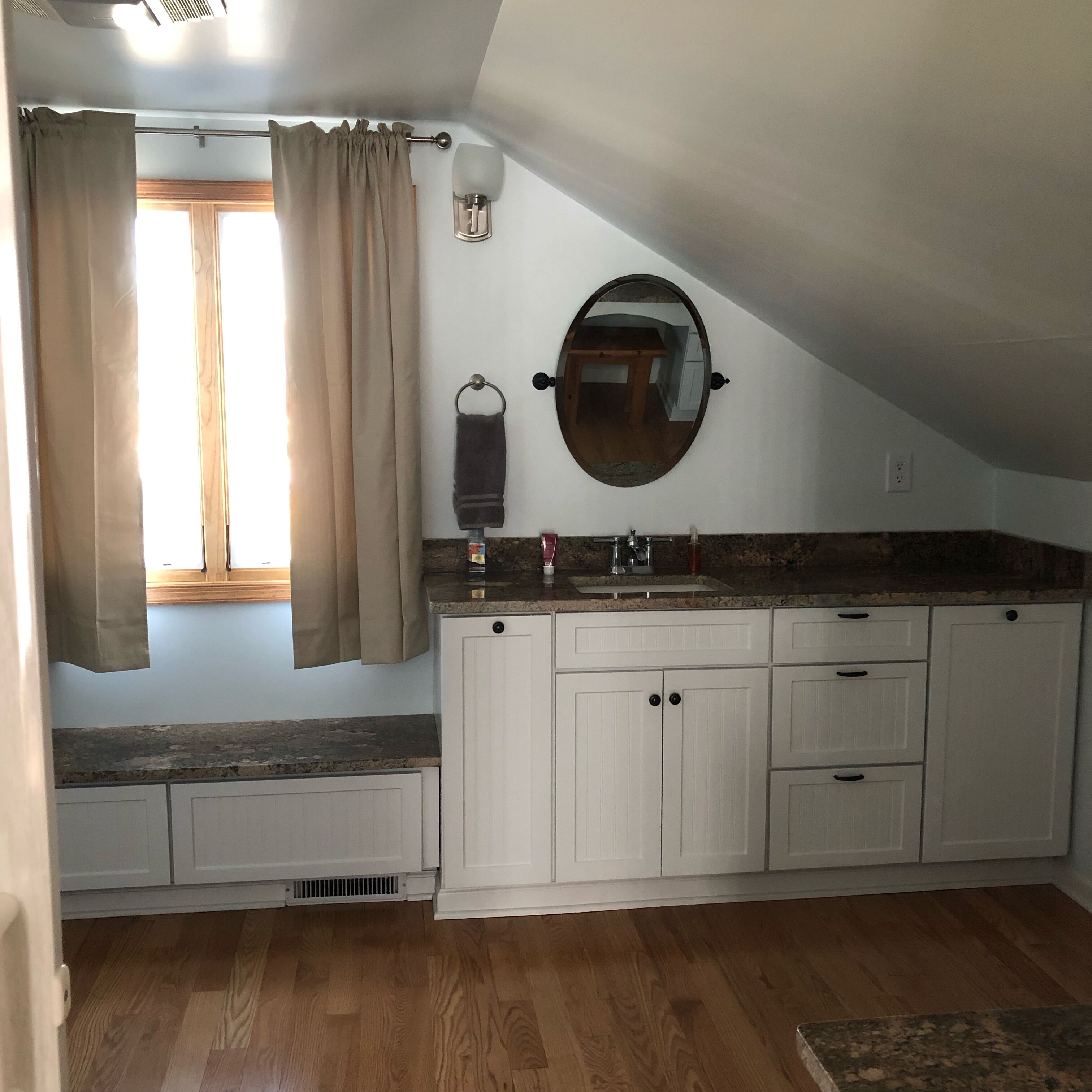A kitchen with white cabinets, a granite countertop, a window with beige curtains, and a wall-mounted mirror.